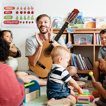 Ein Erzieher sitzt mit seiner Gitarre im Schneidersitz auf dem Boden und klatscht während die Kinder im Kreis um ihn herumsitzen und ihm zuhören